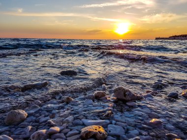 Romantic sunset by a stony seashore. The sun sets over the horizon. The sun beams reflecting in the calm sea waters. Stony shore is washed by the gentle waves. The sky is turning yellow and orange.