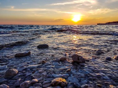 Romantic sunset by a stony seashore. The sun sets over the horizon. The sun beams reflecting in the calm sea waters. Stony shore is washed by the gentle waves. The sky is turning yellow and orange.