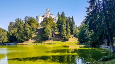 A view on Trakoscan castle from the lake side. Castle is located on a small hill and well hidden in between the trees. The lake in front of the castle is really green. Clear and bright day.
