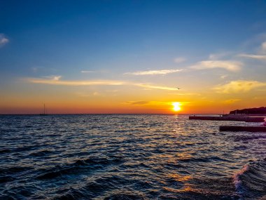 Romantic sunset by a stony seashore. The sun sets over the horizon. The sun beams reflecting in the calm sea waters. Stony shore is washed by the gentle waves. The sky is turning yellow and orange.