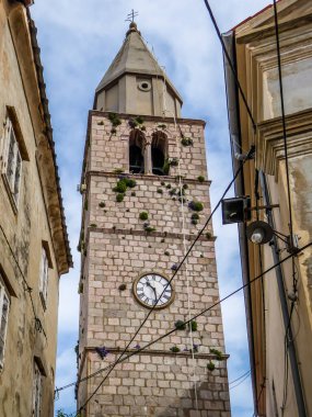 Bell tower located in the middle of an archival city. Tower is towering high above other buildings. Captured from the bottom. All the buildings are coloured beige. Tower overgrown with plants, flowers