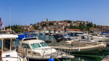 A view on the harbour in Vrsar. There are a lot of boats anchored to the shore. In the back, there is the city center, with a tall church tower. Clear and sunny day. Busy day in the harbour.