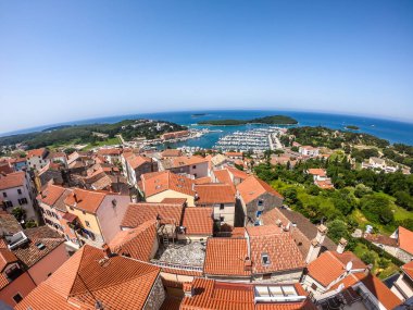 A remote view on the harbour from a hill. Rows of docks waiting for the boats and yachts to anchor there. Lots of houses surrounding the port. In the back there are few islands. Lush green plants