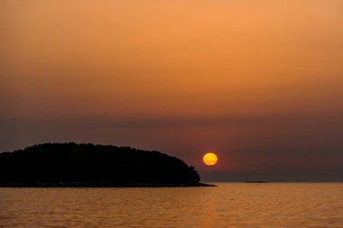 Romantic sunset by a beach. The sun sets over the horizon. The sun beams reflecting in the calm sea waters. There is an island on the side. Few birds flying around. The sky turns yellow and orange