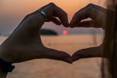 A heart formed from hands, and framing the setting sun in it. The sun sets over the horizon. The sun beams reflecting in the calm sea waters. The sky turns yellow and orange. An island on the side