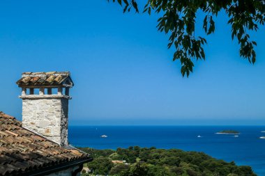 A view on the sea from behind a house. A chimney and a little bit of the roof visible on the side. Tree branches hanging from the top. There are few boats crossing the sea. Clear and sunny day.