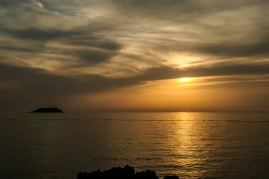 Romantic sunset by a beach. The sun sets over the horizon. The sun beams reflecting in the calm sea waters. There is an island on the side. Few birds flying around. The sky turns yellow and orange