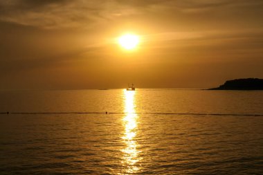 Romantic sunset by a beach. The sun sets over the horizon. The sun beams reflecting in the calm sea waters. There is an island on the side. Few birds flying around. The sky turns yellow and orange