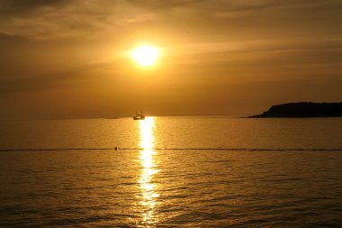 Romantic sunset by a beach. The sun sets over the horizon. The sun beams reflecting in the calm sea waters. There is an island on the side. Few birds flying around. The sky turns yellow and orange