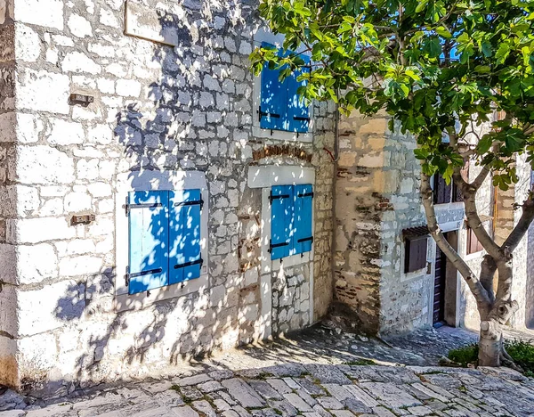 A part of little stony house with blue and brown shutters. The view on the house is disturbed by a tree, growing next to it. Tree casts a shadow on the stony wall. Mediterranean culture.