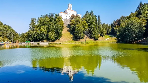 A view on Trakoscan castle from the lake side. Castle is located on a small hill and well hidden in between the trees. The lake in front of the castle is really green. Clear and bright day.