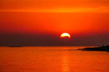 Romantic sunset by a beach. The sun sets over the horizon. The sun beams reflecting in the calm sea waters. There is an island on the side. Few birds flying around. The sky turns yellow and orange