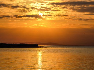 Sunset over the lake. Sun goes down behind the clouds. Sunbeams reflecting in the calm surface of the lake. A small island in the middle of the lake. Neusiedlersee in Austria. Pebbles beach.