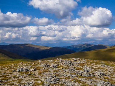 Hiking trails in the high mountain. Green grass, bushes and stones cover the slopes. Endless chains of mountains visible. Thick clouds on the sky. Solo walk allowing for reverie and reflection.
