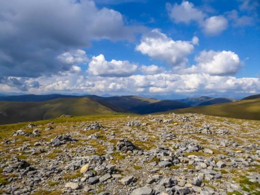 Hiking trails in the high mountain. Green grass, bushes and stones cover the slopes. Endless chains of mountains visible. Thick clouds on the sky. Solo walk allowing for reverie and reflection.
