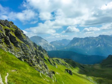 A stunning view on Alpine valley. Hiking trails in the high mountain. Green grass and bushes cover the slopes. Endless chains of mountains visible. Partially cloudy day. Spring in the mountains.