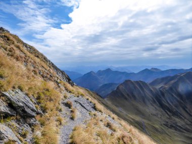 A stunning view on Alpine valley. The trail is overgrown with golden grass. Hiking trails in the high mountain. Endless chains of mountains visible. Partially cloudy day. Autumn in the mountains.