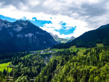A stunning view on Alpine valley. Hiking trails in the high mountain. Green grass and bushes cover the slopes. There is a small settlement in the bottom of the valley. Spring in the mountains.