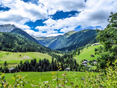 A stunning view on Alpine valley. Hiking trails in the high mountain. Green grass and bushes cover the slopes. There is a small settlement in the bottom of the valley. Spring in the mountains.