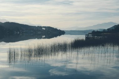 A view on a lake and Alps in the back. The calm surface of the lake is reflecting the mountains, sunbeams and clouds. Clear and sunny day. Calm and relaxed feeling.