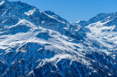 A rescue helicopter in the high mountains. Tall Alps surrounding the vehicle that is getting ready to land. Mountains slopes are covered with thick snow. Dangerous rescue mission.