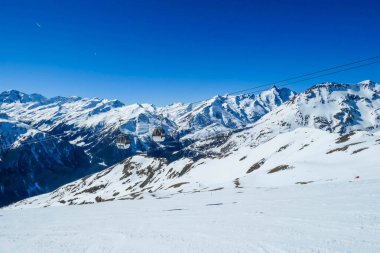 Two gondolas going up and down above the snow caped mountains. Gondolas are moving on thick ropes hanging very high. In the back there are tall Alps. Ski resort in Heiligenblut, Austria.