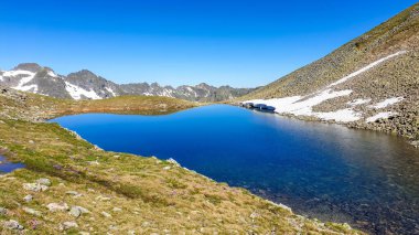Clear, navy blue lake hiding between tall mountain peaks. Some of the slopes are still covered with snow. In the back here is another mountain range. Clear and bright day. Perfect condition for hiking