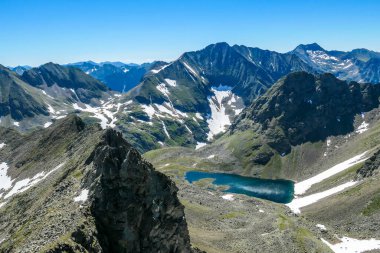A distance view on a clear, navy blue lake hiding between tall mountain peaks. Some of the slopes are still covered with snow. In the back here is another mountain range. Clear and bright day.