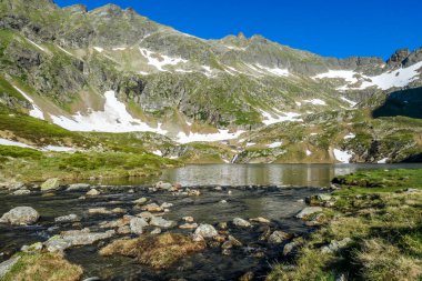 View on an icy-cold alpine lake. There is a lot of snow around on mountain's slopes. Spring is slowly coming into the valley. Calm surface of the lake reflects the mountains. Schladming Alps, Austria