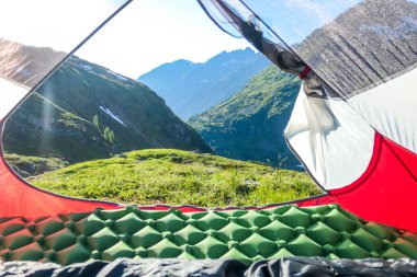 A view through an unfolded tent on a meadow in high Alps. Slopes of the mountains partially covered with snow. Wild camping in Austria. High mountains around. Spring in alpine valleys.