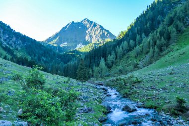 A view on Schladming Alps, partially still covered with snow. A small stream flowing between the stones. Spring slowly reaching the tallest parts of the mountains. First sunbeams reaching the peak.
