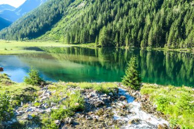 A calm alpine lake. The lake is surrounded with tall mountains. The surface of the lake is calm, it reflects the mountains and sky. Clear and sunny day. Schladming region, Austria
