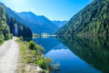 A calm alpine lake. The lake is surrounded with tall mountains. The surface of the lake is calm, it reflects the mountains and sky. Clear and sunny day. Schladming region, Austria