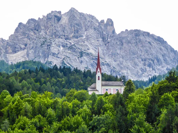 A church popping out of the forest, with a tall, sharp and rocky mountains behind. The church is build on a rock, making it taller than surrounding trees. Massive Alps in the back. Clear day.