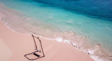 A swing placed on the seashore of Pink Beach, Lombok, Indonesia. The swing has very simple wood construction. Waves gently wash the pillars of it. In the back there are few boats. Drone capture.