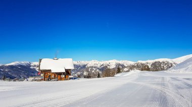 A chalet in Bad Kleinkirchheim, Austria. The little cottage is located next to a ski slope, surrounded by thick snow. The slope is perfectly gravelled. There are snow caped mountains in the back.