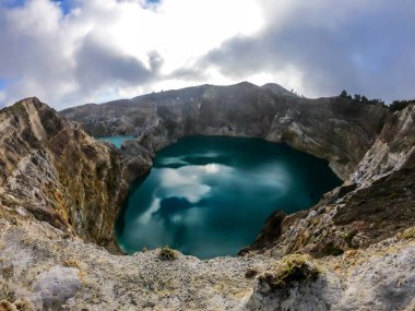 View on the Kelimutu volcanic crater lakes in Flores, Indonesia. Lakes are shining with many shades of turquoise and blue. Sun shines through clouds. Barren and sharp slopes of the volcanic crater