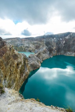 View on the Kelimutu volcanic crater lakes in Flores, Indonesia. Lakes are shining with many shades of turquoise and blue. Sun shines through clouds. Barren and sharp slopes of the volcanic crater