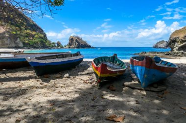 A row of colorful boats parked on a shore of idyllic Koka Beach in Flores, Indonesia. The boats are lined up under the trees, in the shade. There are some cliffs in the back. Hidden gem of Indonesia.