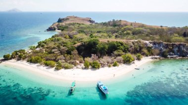 A drone shot of a paradise island with some boats anchored around in Komodo National Park, Flores, Indonesia. Green middle part of the island turns into white sand beach and further into turquoise sea