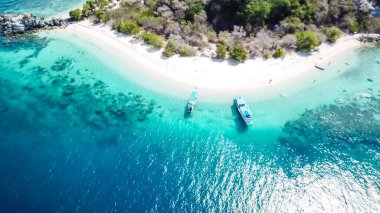 A drone shot of a paradise island with some boats anchored around in Komodo National Park, Flores, Indonesia. Green middle part of the island turns into white sand beach and further into turquoise sea