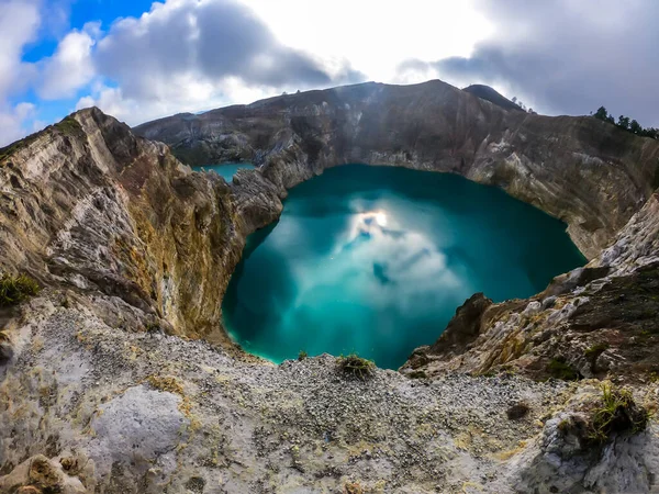 View on the Kelimutu volcanic crater lakes in Flores, Indonesia. Lakes are shining with many shades of turquoise and blue. Sun shines through clouds. Barren and sharp slopes of the volcanic crater