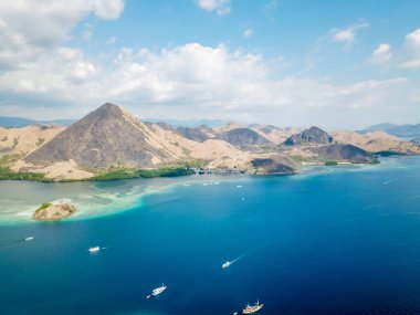 A drone shot of a paradise island with some boats anchored around in Komodo National Park, Flores, Indonesia. Brownish island turns into white sand beach and further into turquoise and navy sea.