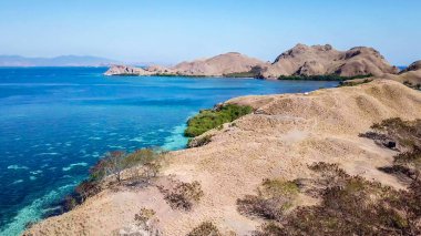 A drone shot of a paradise island in Komodo National Park, Flores, Indonesia. Brownish island turns into white sand beach and further into turquoise and navy blue sea. Steep slopes of the island