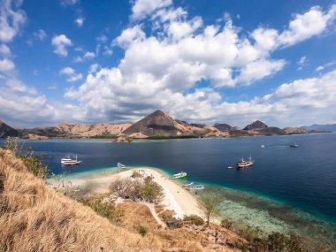 A view from the top of a cliff of Kelor Island on the bay and white sand beach, Komodo National Park, Indonesia. There is another island in the back. There are boats anchored to the shore. Happiness
