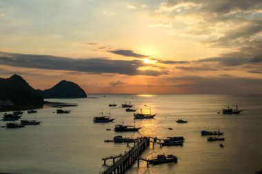 View on a harbour in Labuan Bajo, Flores, Indonesia during the sunset. Sun sets behind the horizon line. Many boats are anchored to shore. There are few islands in the back. Golden hour in a harbour