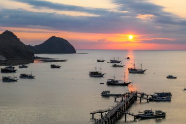 View on a harbour in Labuan Bajo, Flores, Indonesia during the sunset. Sun sets behind the horizon line. Many boats are anchored to shore. There are few islands in the back. Golden hour in a harbour clipart
