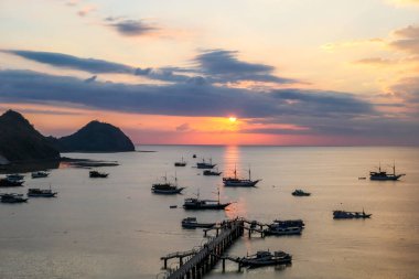 View on a harbour in Labuan Bajo, Flores, Indonesia during the sunset. Sun sets behind the horizon line. Many boats are anchored to shore. There are few islands in the back. Golden hour in a harbour