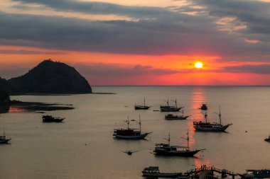 View on a harbour in Labuan Bajo, Flores, Indonesia during the sunset. Sun sets behind the horizon line. Many boats are anchored to shore. There are few islands in the back. Golden hour in a harbour