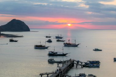 View on a harbour in Labuan Bajo, Flores, Indonesia during the sunset. Sun sets behind the horizon line. Many boats are anchored to shore. There are few islands in the back. Golden hour in a harbour